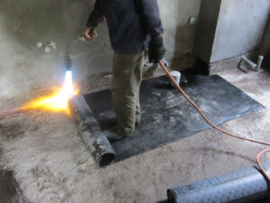 Hình ảnh một thợ thi công đang sử dụng đèn khò để làm nóng và dán màng chống thấm khò nóng lên sàn hoặc tường nhà vệ sinh. Ảnh có thể chụp cận cảnh quá trình thi công, cho thấy lớp màng đang tan chảy và bám dính vào bề mặt