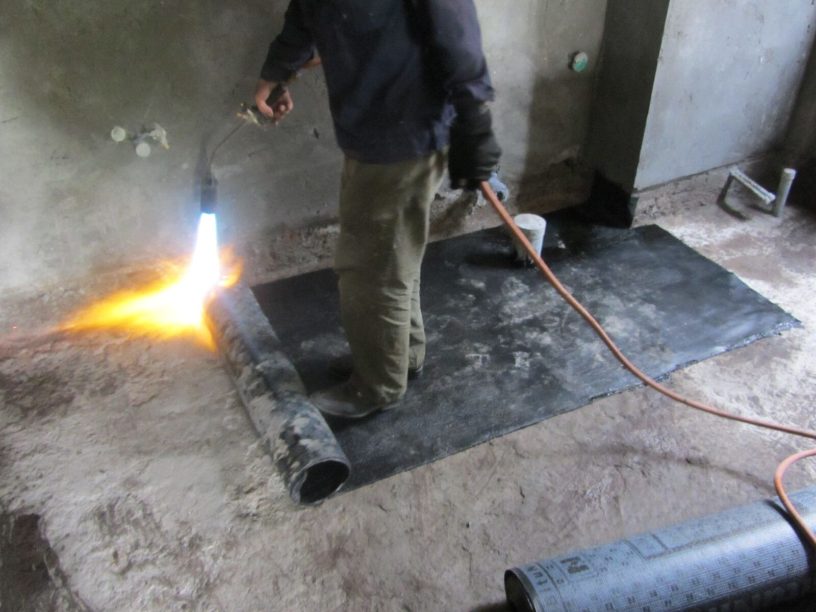 Hình ảnh một thợ thi công đang sử dụng đèn khò để làm nóng và dán màng chống thấm khò nóng lên sàn hoặc tường nhà vệ sinh. Ảnh có thể chụp cận cảnh quá trình thi công, cho thấy lớp màng đang tan chảy và bám dính vào bề mặt