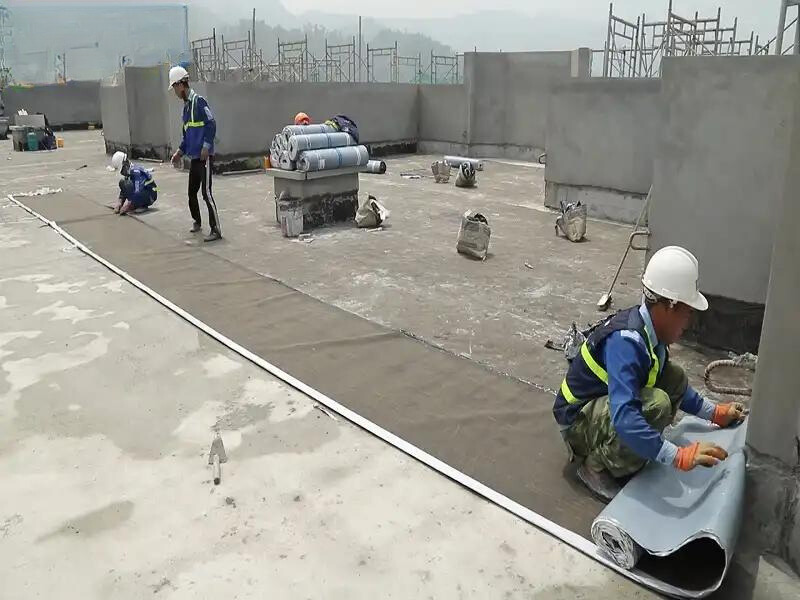 Một công nhân đang làm việc trên mái nhà, kiểm tra và lắp đặt màng chống thấm bitum. Hình ảnh cho thấy tầm quan trọng của việc bảo trì và cải tạo hệ thống chống thấm để ngăn ngừa thấm nước từ trên cao.