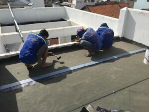 Hình ảnh các kỹ thuật viên đang thi công màng chống thấm Bitum trên bề mặt mái nhà. Họ sử dụng các dụng cụ chuyên dụng để dán màng lên bề mặt, thể hiện tính chuyên nghiệp và sự chắc chắn trong quy trình chống thấm