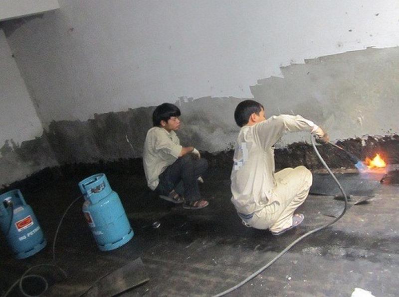 Kỹ thuật viên đang thi công màng chống thấm bitum trên tường tầng hầm, minh họa quy trình bảo vệ kết cấu khỏi nước ngầm.