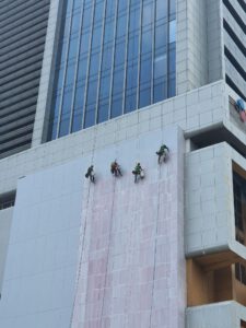 Hình ảnh một nhóm công nhân đang thi công chống thấm trên tường ngoài trời của một tòa nhà cao tầng. Công nhân đang sử dụng các dụng cụ như chổi quét sơn hoặc máy phun màng chống thấm. Bề mặt tường đang được xử lý, thể hiện rõ quy trình làm việc chuyên nghiệp và sự cẩn thận trong thi công