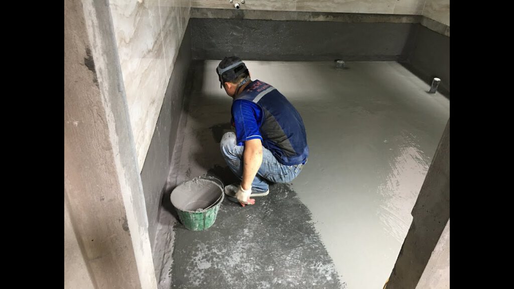 Công nhân đang sử dụng màng chống thấm hoặc sơn chống thấm trên bề mặt sàn nhà vệ sinh, với các công cụ như chổi quét hoặc con lăn.