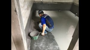 Công nhân đang sử dụng màng chống thấm hoặc sơn chống thấm trên bề mặt sàn nhà vệ sinh, với các công cụ như chổi quét hoặc con lăn.