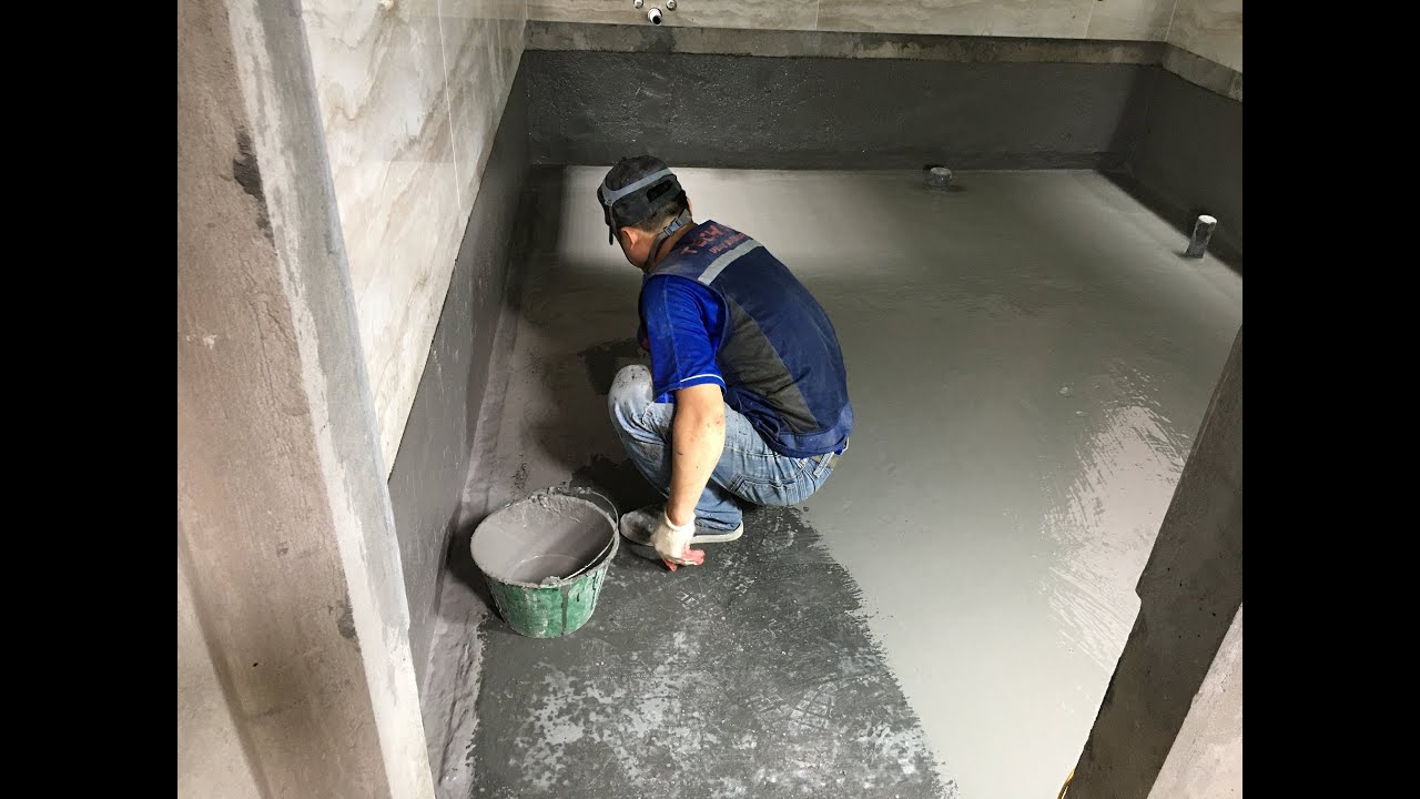 Công nhân đang sử dụng màng chống thấm hoặc sơn chống thấm trên bề mặt sàn nhà vệ sinh, với các công cụ như chổi quét hoặc con lăn.